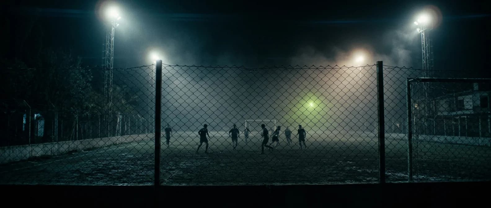 Un partido de fútbol 7 de noche visto a través del alambrado, bajo los reflectores y la bruma